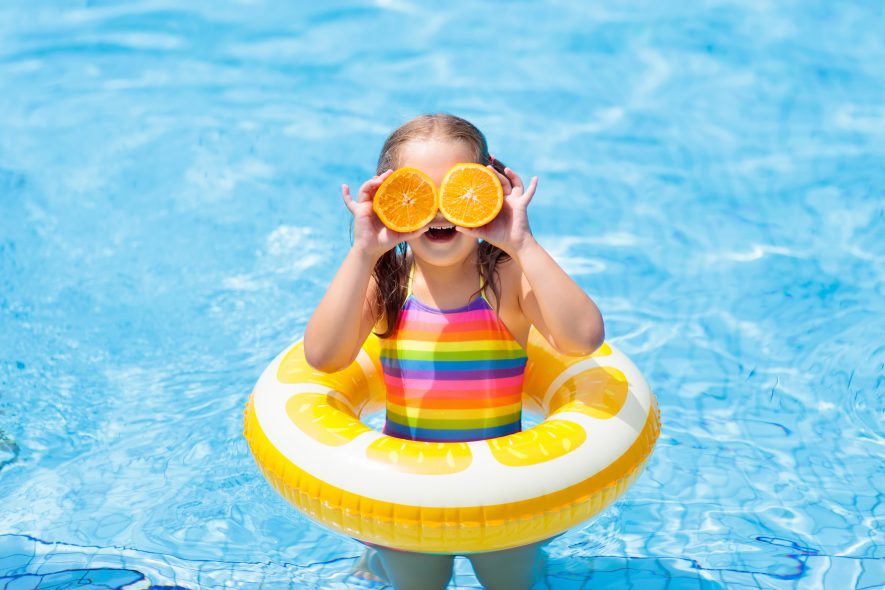 Little girl in swimming pool with inflatable toy ring eating orange. Kids swim on summer vacation. Tropical fruit and healthy snack. Swim aids for child.  Kid on colorful float. Beach and water fun.